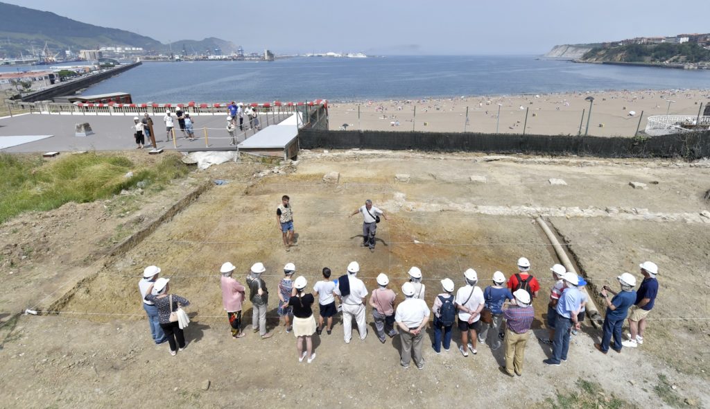 Un grupo de visitantes atienden las explicaciones del arqueólogo, al pie del yacimiento de Punta Begoña. Vista aérea hacia la bahía de El Abra